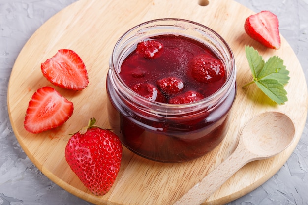 Strawberry jam in a glass jar with berries and leaves on gray concrete 