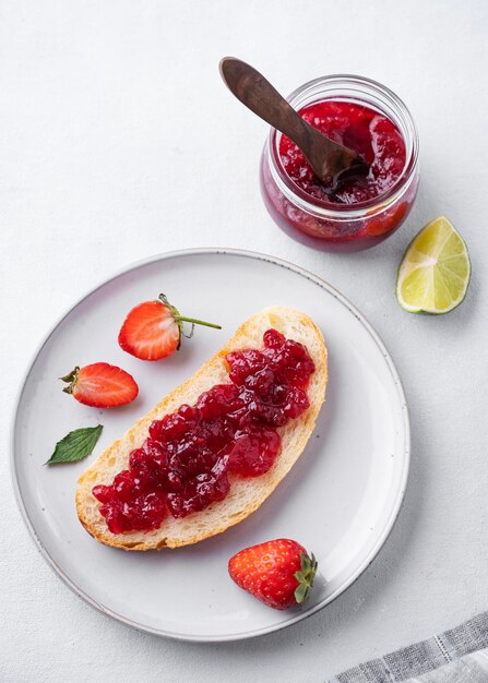 Strawberry jam on a piece of bread on a plate on a blue background with fresh berries and jar with jam