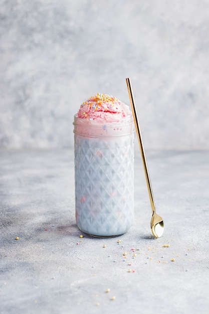 Strawberry milkshake with strawberry ice cream and blue spirulina milk and pastel colorful confetty topping with gold spoon on gray stone wall. close up