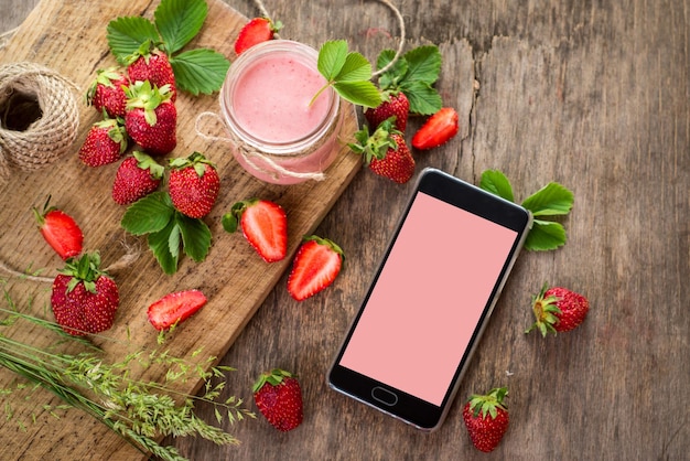 Strawberry smoothies in glass with many strawberries and electronic gadget on wooden background. Top view. Copy space