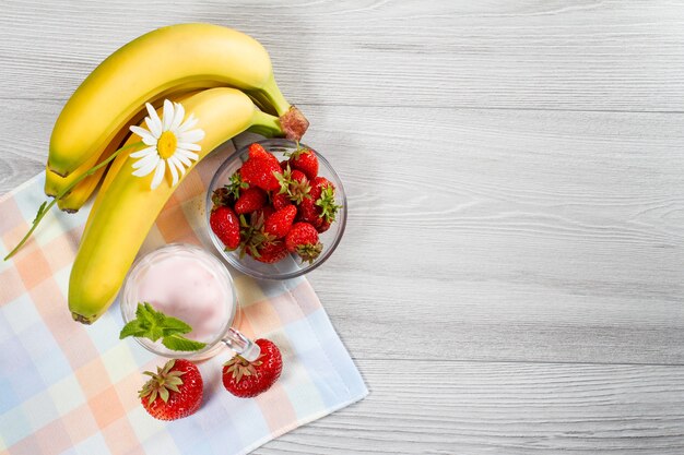 Foto yogurt alla fragola in un bicchiere con fragole fresche, banana, camomilla su un tavolo di legno con un tovagliolo vista dall'alto