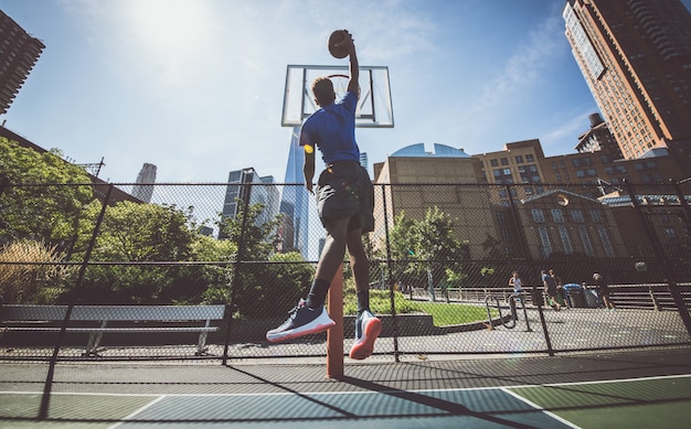 Street basketball athlete performing huge slam dunk on the court, New york buildings