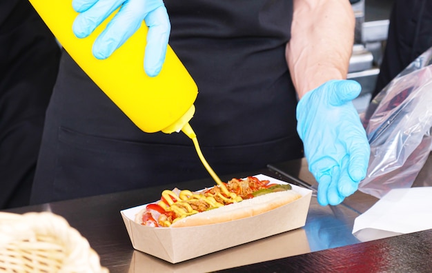 Street food cart worker prepares a hot dog. gloved hands add\
mustard to the hot dog. traditional takeaway, street food,\
snacks.