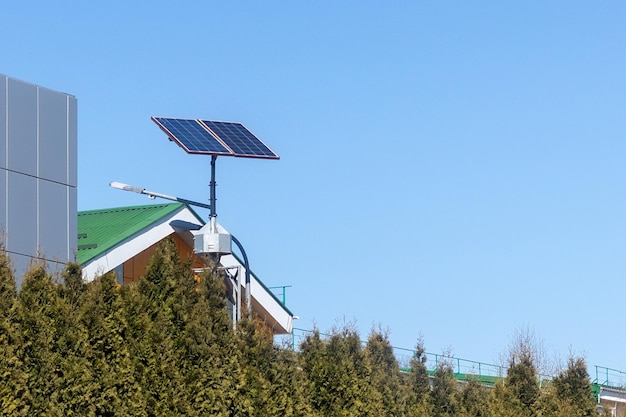 Street lamp with a solar panel behind green trees against a background of roofs and blue sky