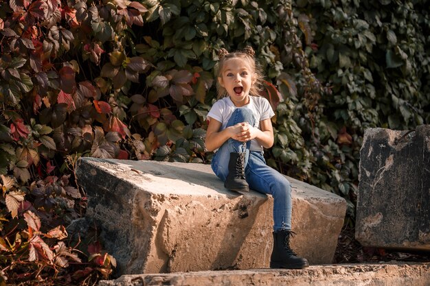 Street photo of a little beautiful girl in jeans and a white t-shirt on a background of concrete slabs and autumn leaves