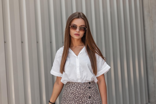 Street portrait of a beautiful fashionable business girl with vintage sunglasses wearing summer casual clothes with a white shirt and skirt in the near a metal wall