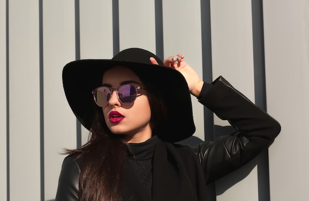 Street portrait of brunette woman in trendy wide brimmed hat and stylish glasses posing in sun light