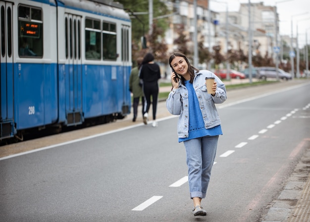 彼女の手にコーヒーと電話で話している陽気な若い女性のストリートポートレート