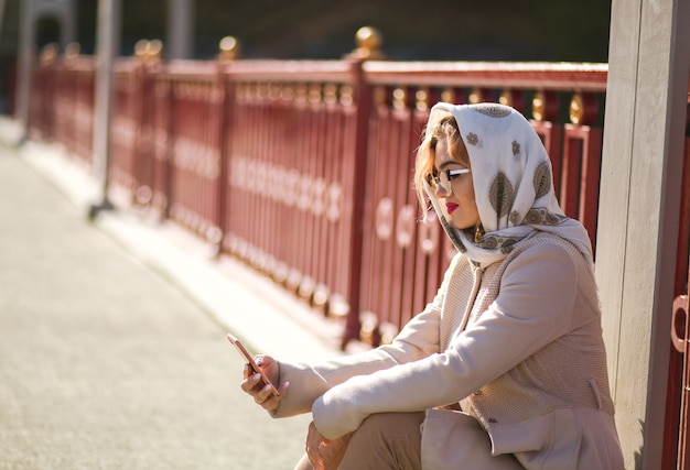 Photo street portrait of elegant blonde model wears beige coat , white shawl and typing a message on her smart phone