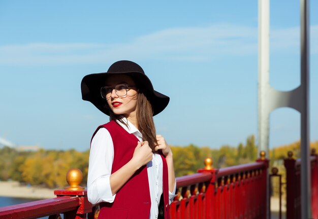 세련된 여성의 거리 초상화는 빨간 의상, 검은 모자, 세련된 안경을 쓰고 화창한 날 포즈를 취합니다. 텍스트를 위한 공간