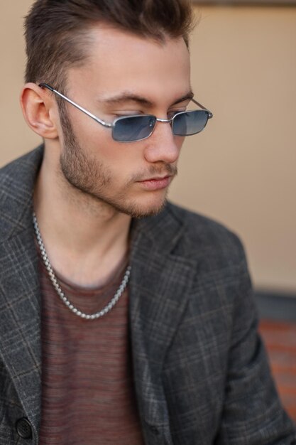 Street portrait of a handsome hipster guy with blue fancy sunglasses in a fashion coat and Tshirt sitting on the street