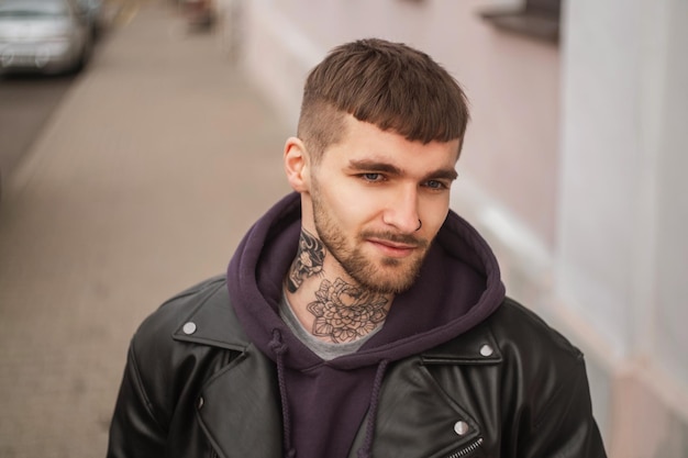 Foto ritratto di strada di un bel ragazzo hipster alla moda con i capelli e la barba con un tatuaggio sul collo in abiti alla moda con giacca e felpa con cappuccio che cammina per la città