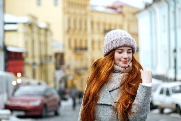 Street portrait of lovely redhead woman with long hair wearing warm winter apparel posing at the street. Space for text