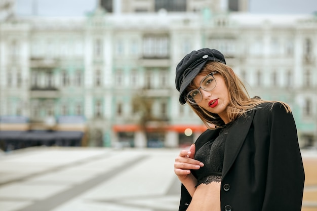 Street portrait of sexy brunette woman with red lips wears stylish cap, glasses and lingerie. Space for text