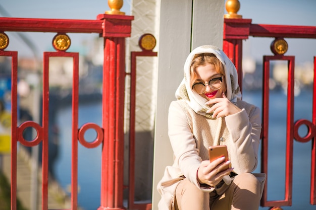 Уличный портрет улыбающейся блондинки носит бежевое пальто, белый платок и печатает сообщение на своем смартфоне