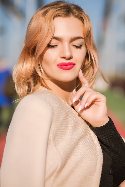 Street shot of beautiful blonde model with bright makeup and perfect skin posing in sun light