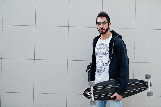 Человек уличного стиля арабский в eyeglasses с longboard представил против серой стены.