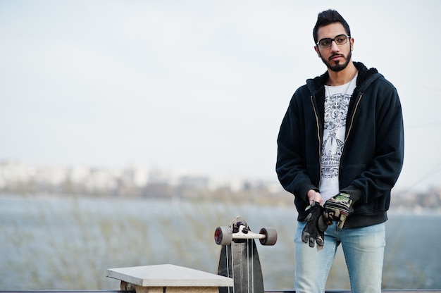 Street style arab man in eyeglasses with longboard posed against river.