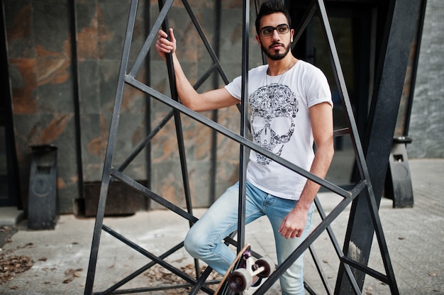 Street style arab man in eyeglasses with longboard posed inside metal pyramid construction.