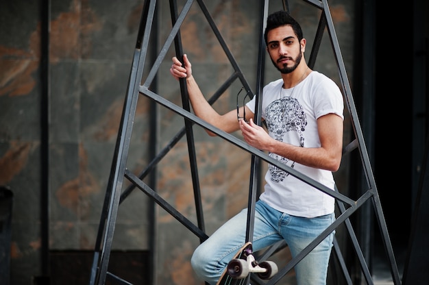 Street style arab man in eyeglasses with longboard posed inside metal pyramid construction.