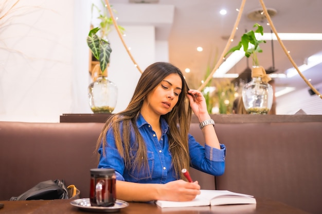 Street Style in the city, a brunette Caucasian girl in a denim shirt having tea in a coffee shop. Writing in a notebook a work for the university