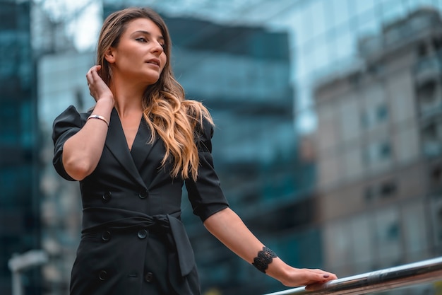 Street style, an enterprising young blonde Caucasian woman in a black suit in the black glass building where she works in the background. Looking to the right and stroking her hair