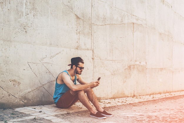 Street Style. Knappe jongeman in koptelefoon die een smartphone vasthoudt en ernaar kijkt terwijl hij buiten zit en tegen de betonnen muur leunt