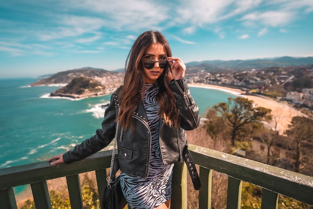 Streetstyle, een jonge brunette in een zwart-witte korte jurk en een leren jas met uitzicht over de stad San Sebastia