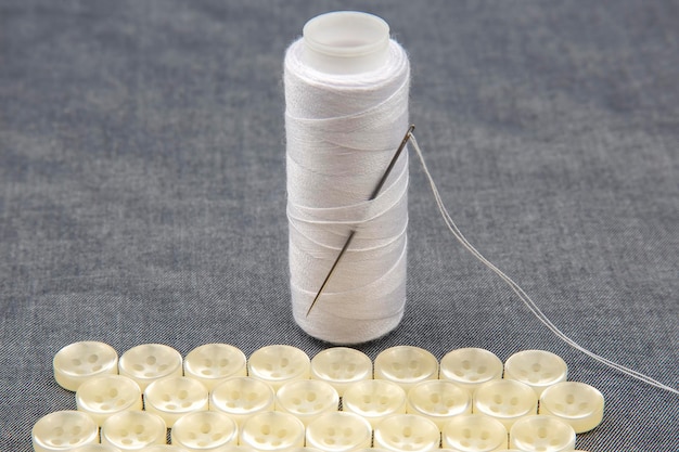 Streng van witte draad en een naainaald. Plastic glanzende knopen voor kleding op een stoffenachtergrond. Mode en kleding. Fabrieksindustrie.