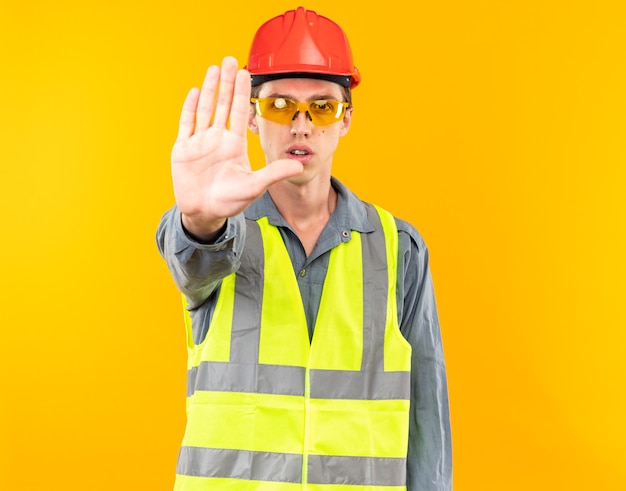 Strenge jonge bouwer man in uniform dragen van een bril met stop gebaar geïsoleerd op gele muur