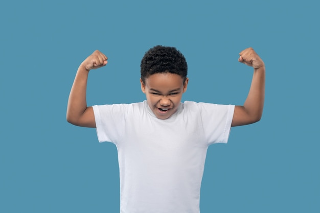 Strength, muscles. Stretching black boy in white tshirt showing strength by straining muscles of his arms on light blue background