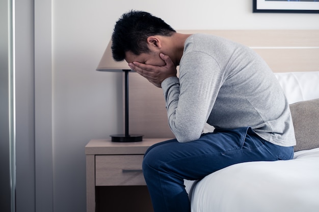 Stress Asian young man sitting alone on bed and crying with tear and cover face by both hands.