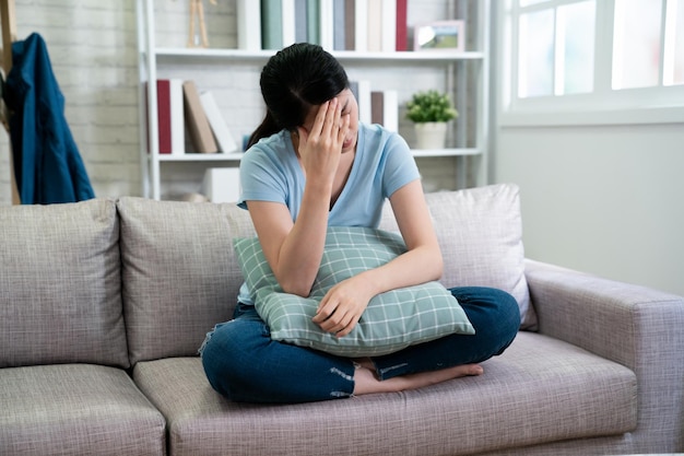 ストレス頭痛ヘルスケアと人々の概念。目を閉じて額に触れる不幸なアジアの女性主婦は、自宅の居心地の良いリビングルームの窓に向かってソファに座って頭痛の病気の病気です。