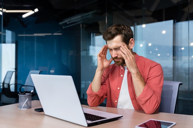 Stress and headache at work a young businessman in a red shirt is sitting at the desk in the office