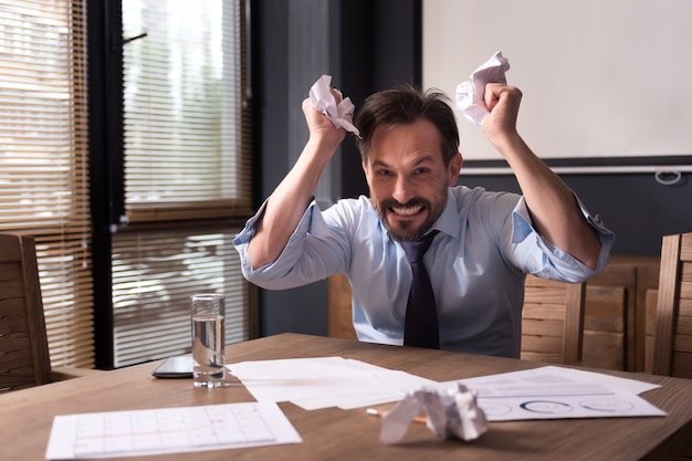 Stress at the workplace. Angry furious hysterical man crumpling paper and grimacing while being at work