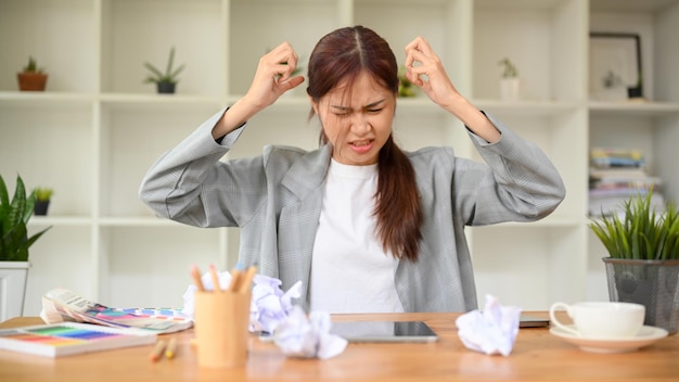 Stressed angry young asian businesswoman sits at office desk\
with crumpled paper