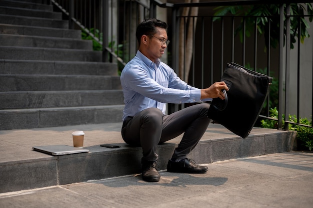 ストレスを感じているアジアのビジネスマンが外の階段に座ってブリーフケースの中に何かを開け見つけています