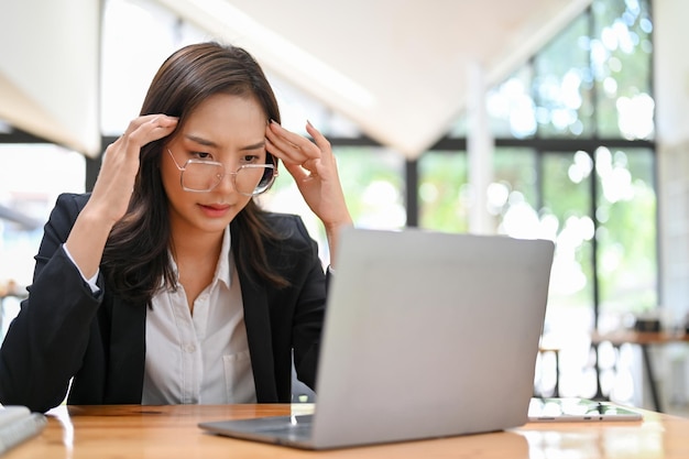 Стрессовая азиатская деловая женщина обдумывает решение кризиса своей компании, работая на ноутбуке