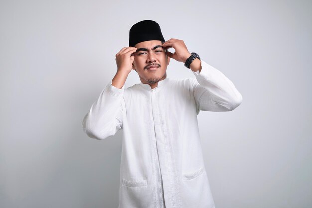 Stressed Asian Muslim man in white clothes and black skull cap touching his temples and having headache