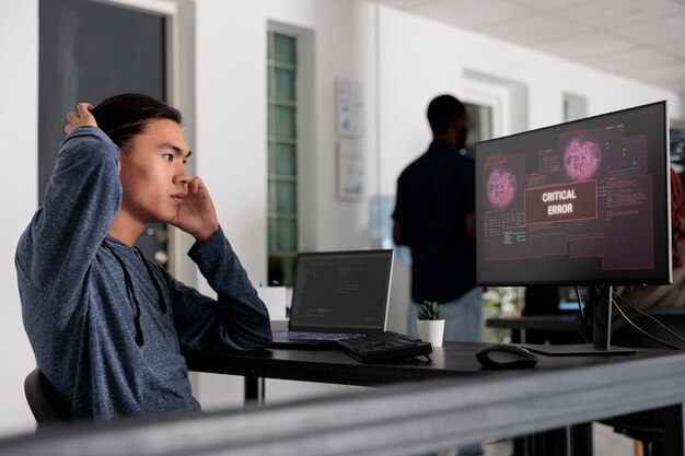stressed asian web developer having security breach alert on computer screen, trying to solve hacking and critical error. programmer using terminal window and having system hacked crash.