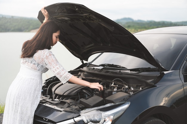 Stressed asian woman open hood, looking a broken car and checking engine on country road.