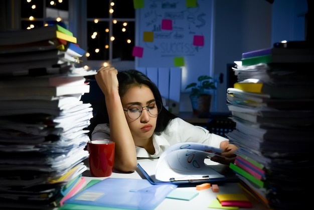 写真 オフィスに座って、テーブルの上の過負荷作業文書で残業しているストレスの多いアジアの女性