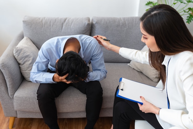 Stressed asian young man patient have life problem sitting on\
sofa with hands holding his head while woman psychiatrist trying to\
console him