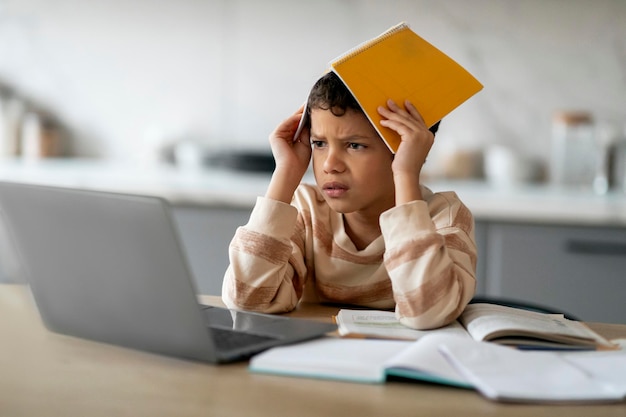 Фото Стрессовый чернокожий мальчик использует ноутбук и покрывает голову рабочей тетрадью дома
