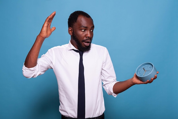 Stressed businessman raising hand to stop alarm clock feeling deadline pressure on blue background. Anxious person in white office shirt and tie looking nervous showing passing time concept.