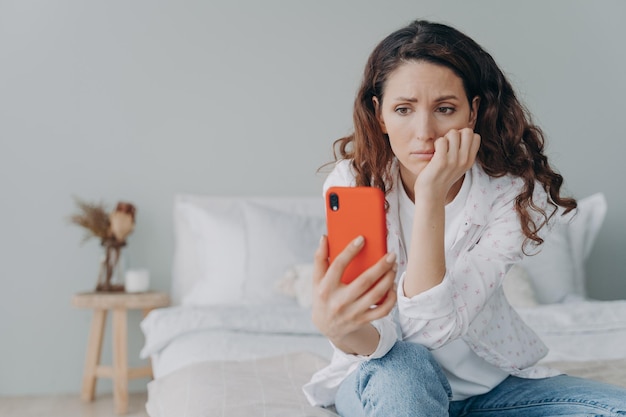 Stressed caucasian woman is doom scrolling Girl reading morning news on smartphone in her bedroom