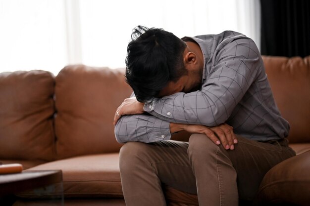 Stressed and depressed millennial Asian man is crying on the sofa in a living room