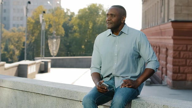 Stressed frustrated elderly African American man of retirement age in casual clothes shirt and jeans sitting outdoor holding phone waiting for client or friend late for meeting anxiously look aside