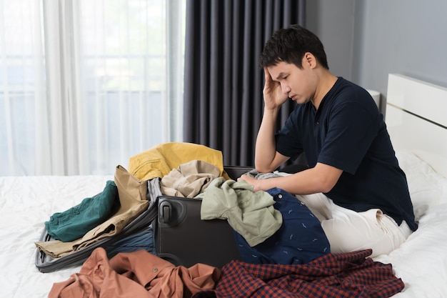 ベッドの休日の旅行の概念でスーツケースに服を詰めるのに問題があるストレスの多い男