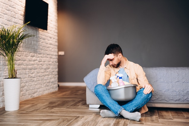 Stressed man looking disappointed before housecleaning, sitting on floor with cleaning supplies